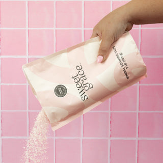 Hand pouring Sweet Grace mineral soaking salts from pink packaging bag against pink tile background.