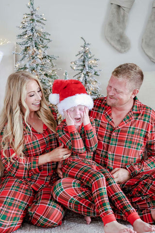Family wearing matching red and green plaid Christmas pajamas sitting in front of holiday trees