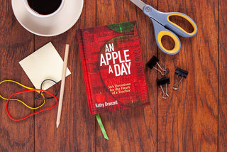 Closed red hardcover An Apple a Day devotional for Christian teachers by Kathy Branzell with white typography on wood