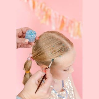 Adult applying blue glitter gel to child's scalp using small brush and clear jar