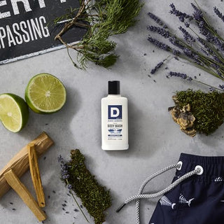 Duke Cannon Midnight Swim Body Wash trial size bottle surrounded by lime, lavender, and driftwood ingredients