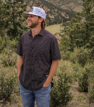 Man wearing a patterned shirt and cap standing in a natural landscape with trees and mountains.
