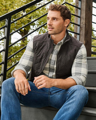 Man wearing dark gray quilted vest over gray plaid flannel shirt and blue jeans