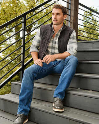 Man wearing dark gray quilted vest over plaid shirt, seated on steps