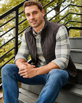 Man wearing dark gray quilted vest over gray plaid flannel shirt and blue jeans
