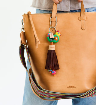 Consuela Charm Lennie colorful bird keychain with brown leather tassel attached to tan leather tote bag