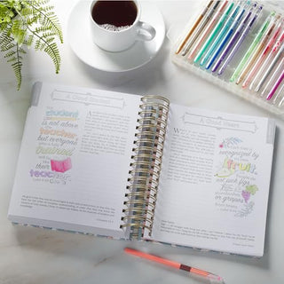 Open spiral-bound coloring devotional with illustrated Bible verses, pens, and coffee nearby