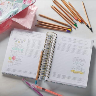 Open spiral-bound coloring devotional journal with colored pencils and pens on white surface