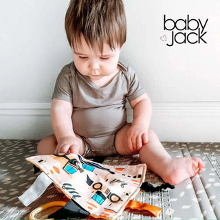 Baby holding beige construction truck sensory lovey with ribbons while sitting on mat