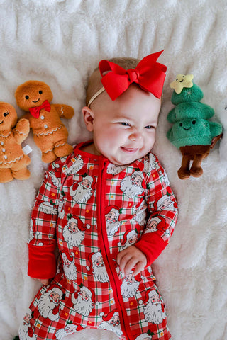 Baby girl in red Santa Claus plaid zip romper with red bow headband, surrounded by Christmas plush toys