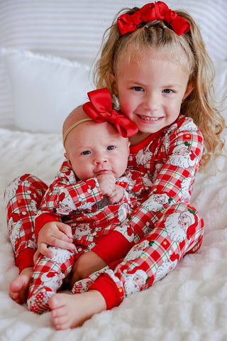Two children wearing matching red plaid Santa Claus print bamboo Christmas pajamas on white bedding.