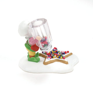 Department 56 elf figurine decorating a star-shaped cookie with colorful sprinkles from a jar