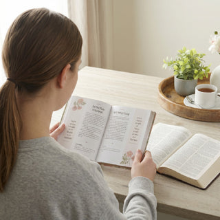 Woman reading a pink floral devotional book at a desk with Bible and tea nearby.