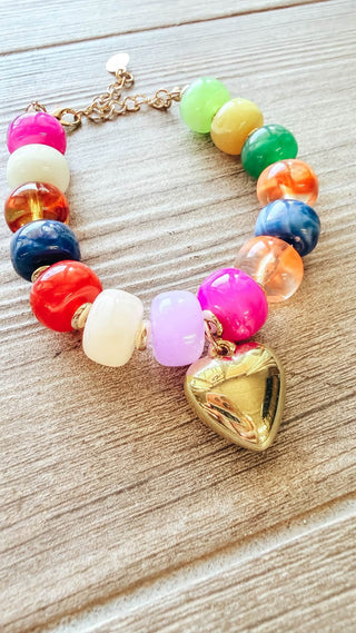 A colorful beaded bracelet with a shiny gold heart charm, displayed on a light brown wooden surface. The bracelet features a variety of vibrant, translucent beads in pink, green, yellow, orange, blue, red, and lavender, connected by a gold-tone chain.