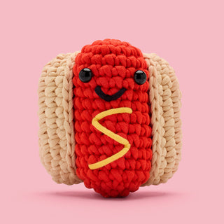 Cute crocheted hot dog amigurumi with smiling face and mustard swirl detail on pink background