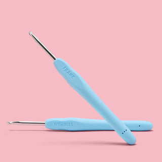 Two light blue ergonomic crochet hooks by The Woobles, one labeled Frank, on pink background