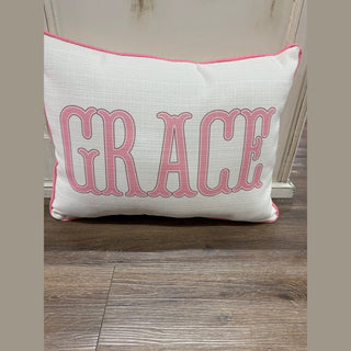 Rectangular off-white polyester name pillow with pink piping and 'GRACE' embroidery for girl's room decor