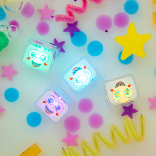 Four Glo Pals light-up sensory cubes with cartoon faces glowing in multicolor light among colorful confetti and stars.