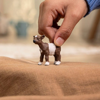 Hand holding a small Schleich Goat Kid figurine with brown and white coloring on burlap surface.