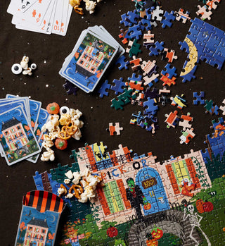 Rifle Paper Co. Haunted House Halloween jigsaw puzzle partially assembled with playing cards and snacks