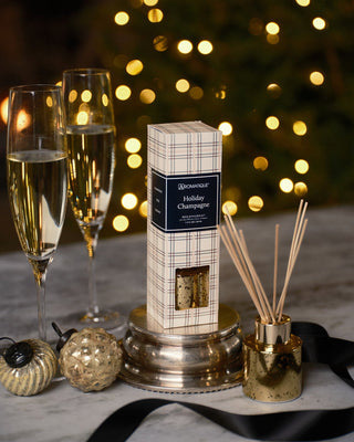 Aromatique Holiday Champagne Mini Reed Diffuser in plaid box with gold vessel and reeds beside champagne flutes