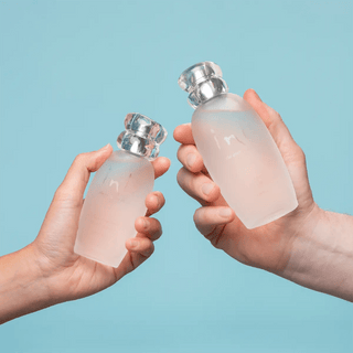 Two hands holding Inis cologne bottles in frosted glass with silver caps against blue background