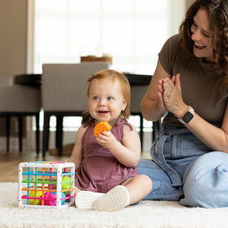 Baby girl happily playing with Fat Brain Toys InnyBin shape sorter toy on white carpet