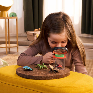 Young girl uses magnifying glass to examine Schleich insect figurines on cork board