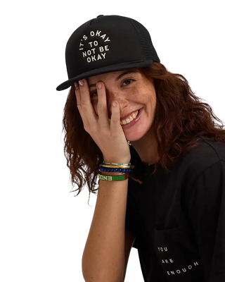 Smiling woman wearing black trucker hat embroidered with It's Okay To Not Be Okay