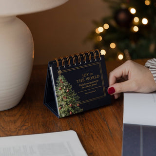 Navy spiral-bound Joy to the World Advent flipbook on wooden desk near Christmas tree