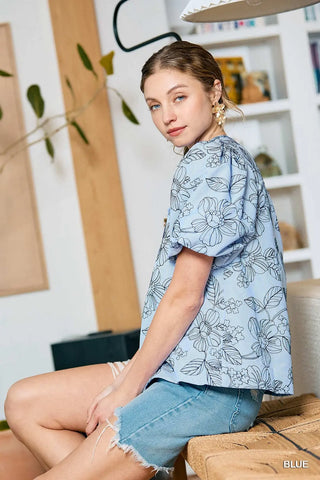 Woman wearing a light blue floral blouse and denim shorts sitting on a couch.