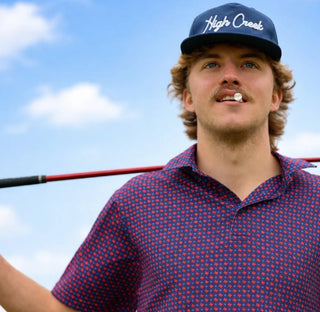 Man wearing a blue cap with 'High Creek' logo and red polka dot shirt holding a golf club against a blue sky.