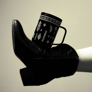 Corkcicle Western Gothic 16oz mug balanced on black leather cowboy boot heel in black and white lifestyle photo