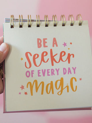 Spiral-bound desk flip calendar displaying 'Be a Seeker of Every Day Magic' quote with colorful typography