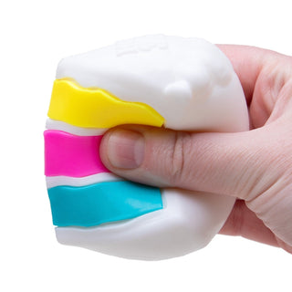 Hand squeezing white Peace O Cake NeeDoh stress ball with colorful yellow, pink, and teal layers