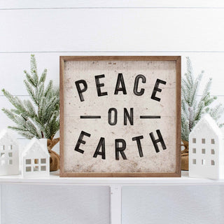 Peace On Earth weathered wood Christmas decor sign on white shelf with miniature paper houses and frosted evergreen branches