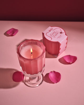 Lit Peony Petals ribbed pink candle in glass vessel beside pink gift box with rose petals
