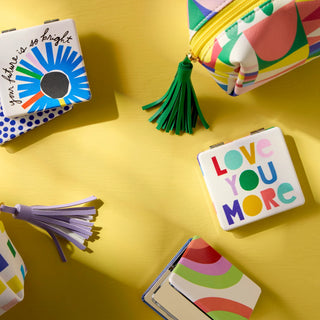Colorful pocket mirrors with uplifting messages displayed on a bright yellow background with accessories.