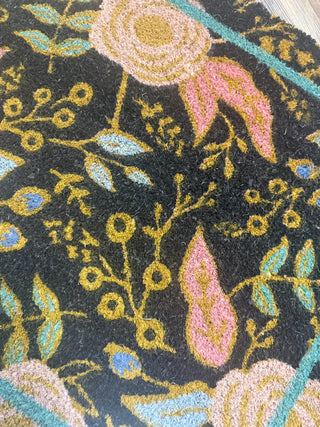 Black coir doormat with gold floral branches, pink paisley teardrops, and teal leaf accents