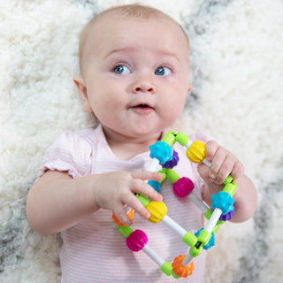 Baby holding colorful Fat Brain Toys Quubi sensory rattle toy while lying on white fluffy surface