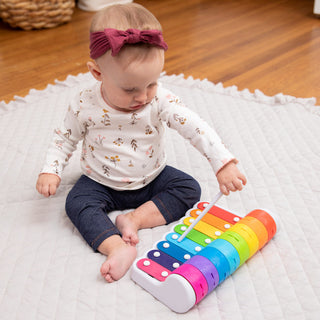 Baby girl playing with Fat Brain Toys Rock N Roller Piano rainbow xylophone on white play mat