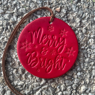 Round red leather ornament embossed with Merry and Bright script and snowflakes on brown cord