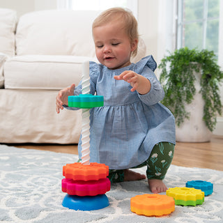 Toddler girl stacking colorful gear-shaped rings onto Fat Brain Toys SpinAgain toy on carpet
