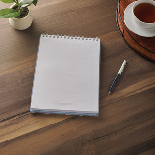 Spiral-bound lined notepad open on wooden desk beside pen and tea cup