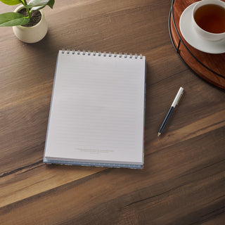 Spiral-bound lined notepad open on a wooden desk beside a pen and tea cup