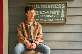 Young man wearing beige Local Boy Outfitters Sportsman's Shacket seated near Wilderness Resort sign