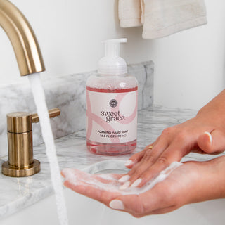 Sweet Grace foaming hand soap bottle on marble sink with lathered hands under running water