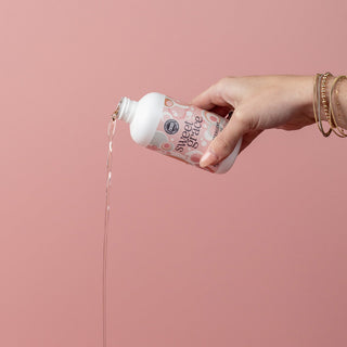 Hand pouring Bridgewater Sweet Grace laundry detergent from pink bottle against pink background.