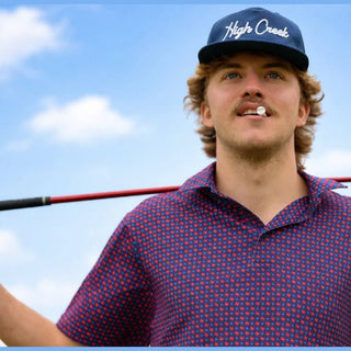 Man wearing a blue cap with 'High Creek' logo and red polka dot shirt holding a golf club against a blue sky.