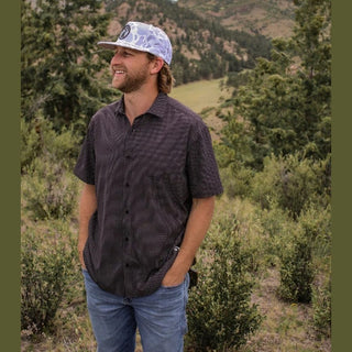 Man wearing a patterned shirt and cap standing in a natural landscape with trees and mountains.